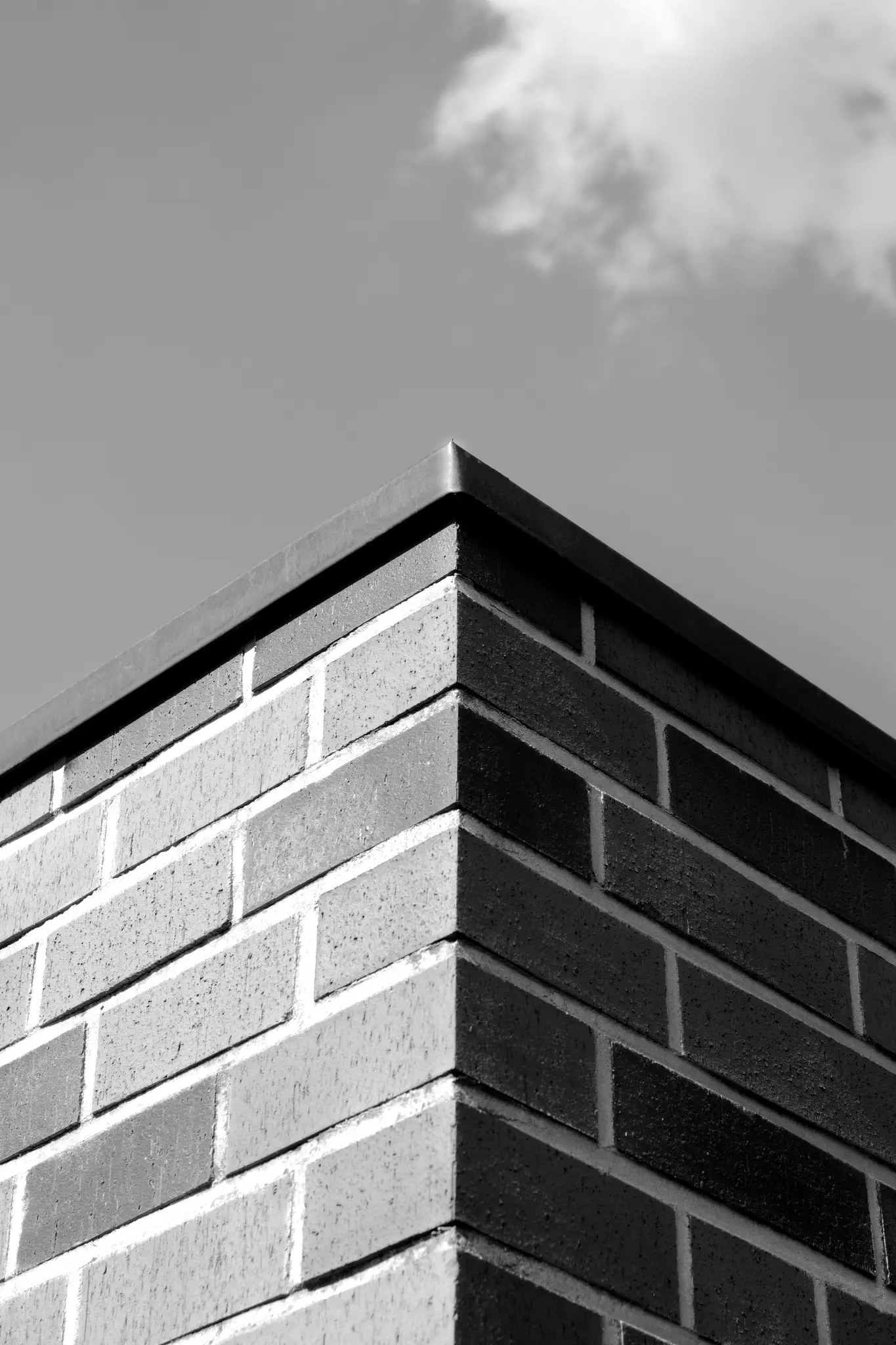 A black and white, low-angle photo of a brick building corner, showing two walls meeting at a sharp angle under a sky with…