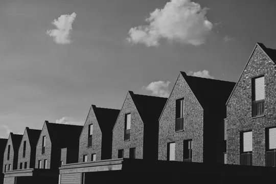 Et sort-hvidt billede viser en række moderne huse med gavle og murstensfacader, under en himmel med skyer.