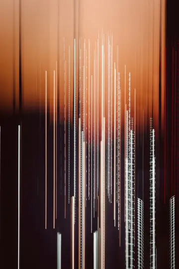 An abstract photo of vertical light trails, predominantly white, against a blurred background of warm orange and dark brown.