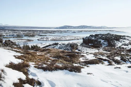 A wide shot of a snowy landscape featuring dark rocks, patches of brown vegetation, a winding body of water, and…