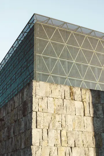 A building corner featuring a rough, light-colored stone wall at the bottom and a modern upper section with a mesh facade.