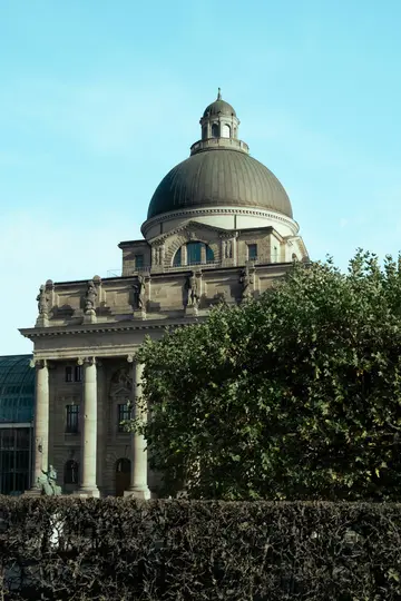 A large, light-colored stone building with a dark dome and classical columns is partially hidden by green trees and a dark…
