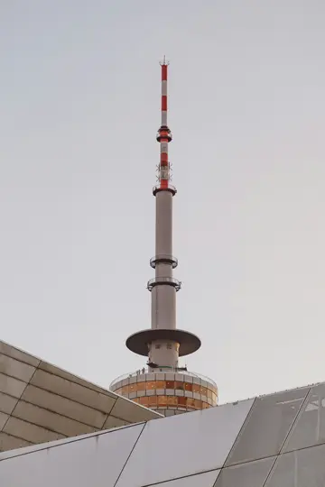 Et højt, gråt kommunikationstårn med en rød og hvid stribet antenne mod en bleg himmel, delvist skjult af kantede grå…