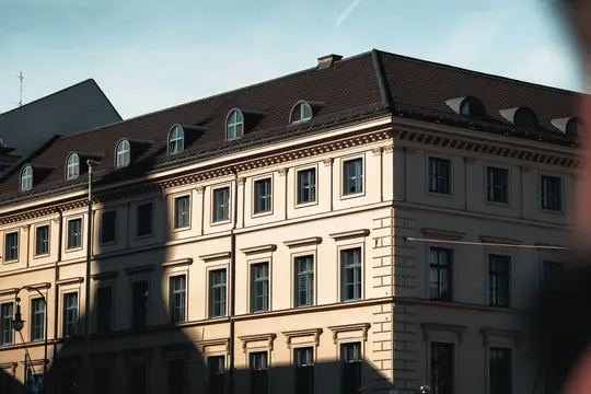 An angled view of a building with a light-colored facade, dark tiled roof, and multiple dormer windows. Strong shadows fall…