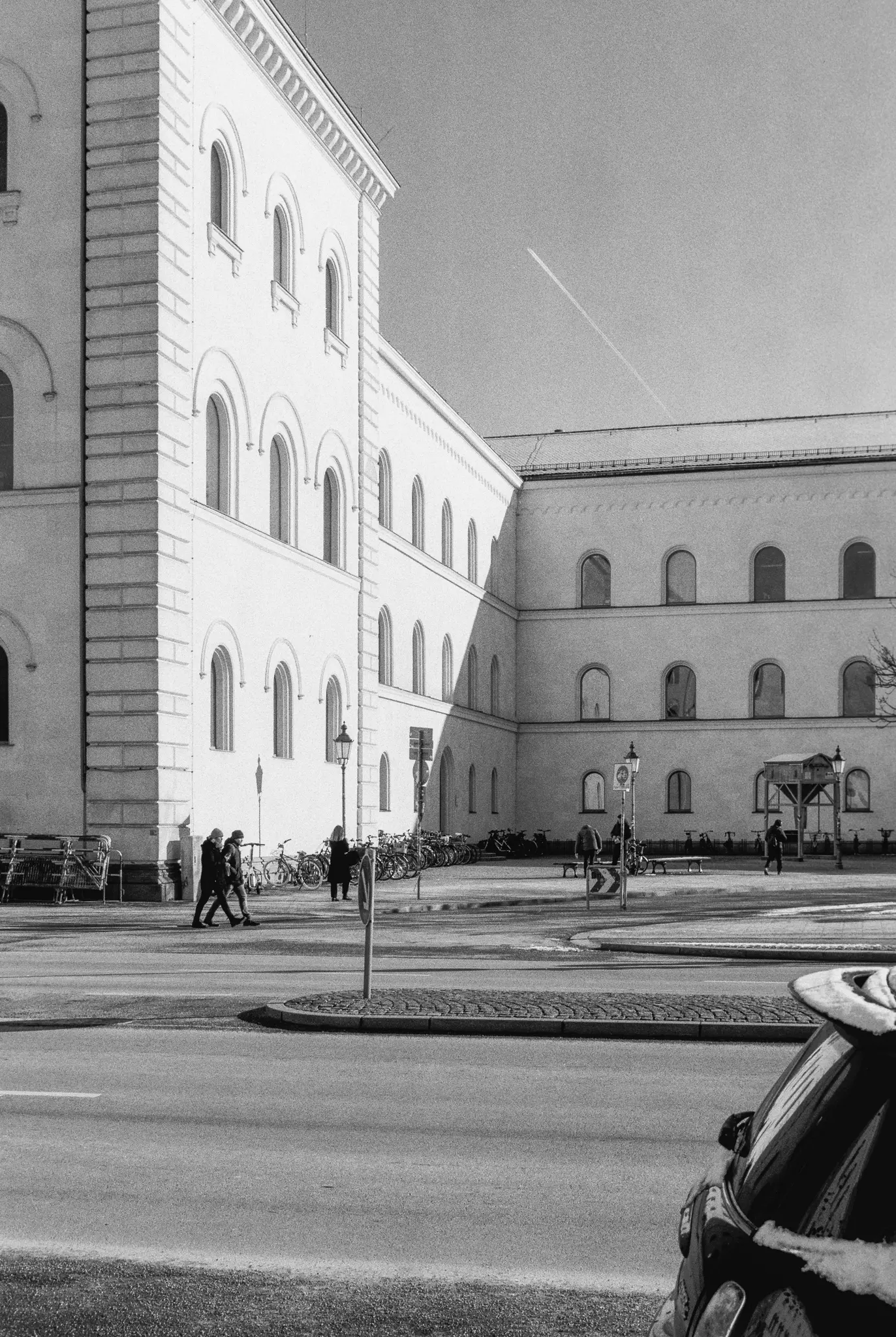 A black and white photo of a street with a large building featuring many arched windows, people walking, and parked bicycles.