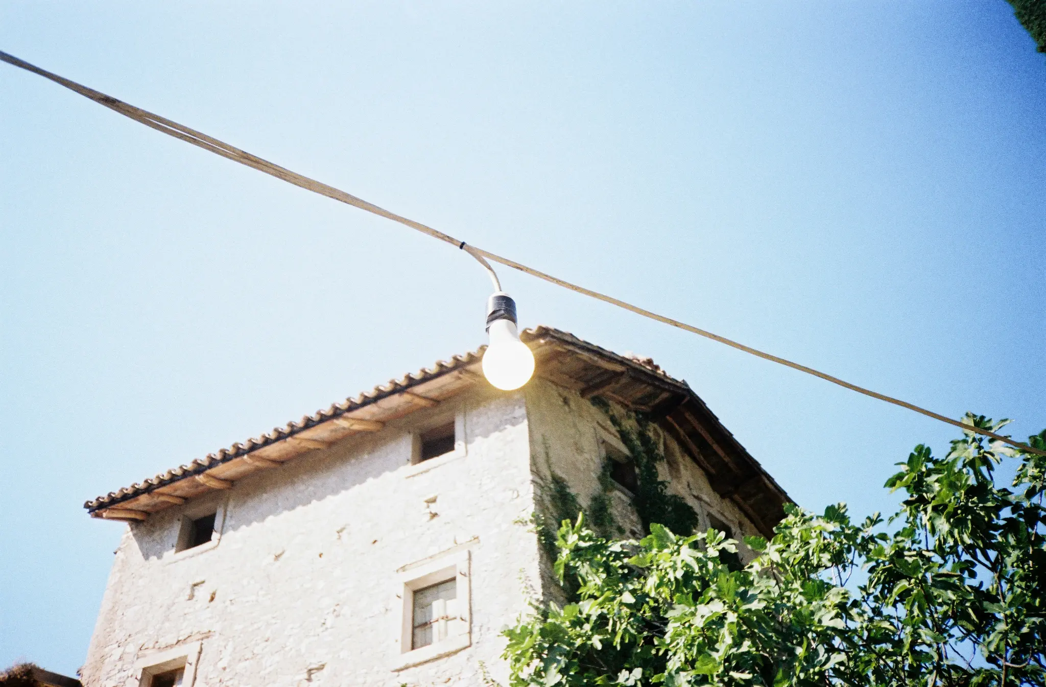 A low-angle photo shows an old, light-colored stone building with a terracotta roof and green vines. An illuminated light…
