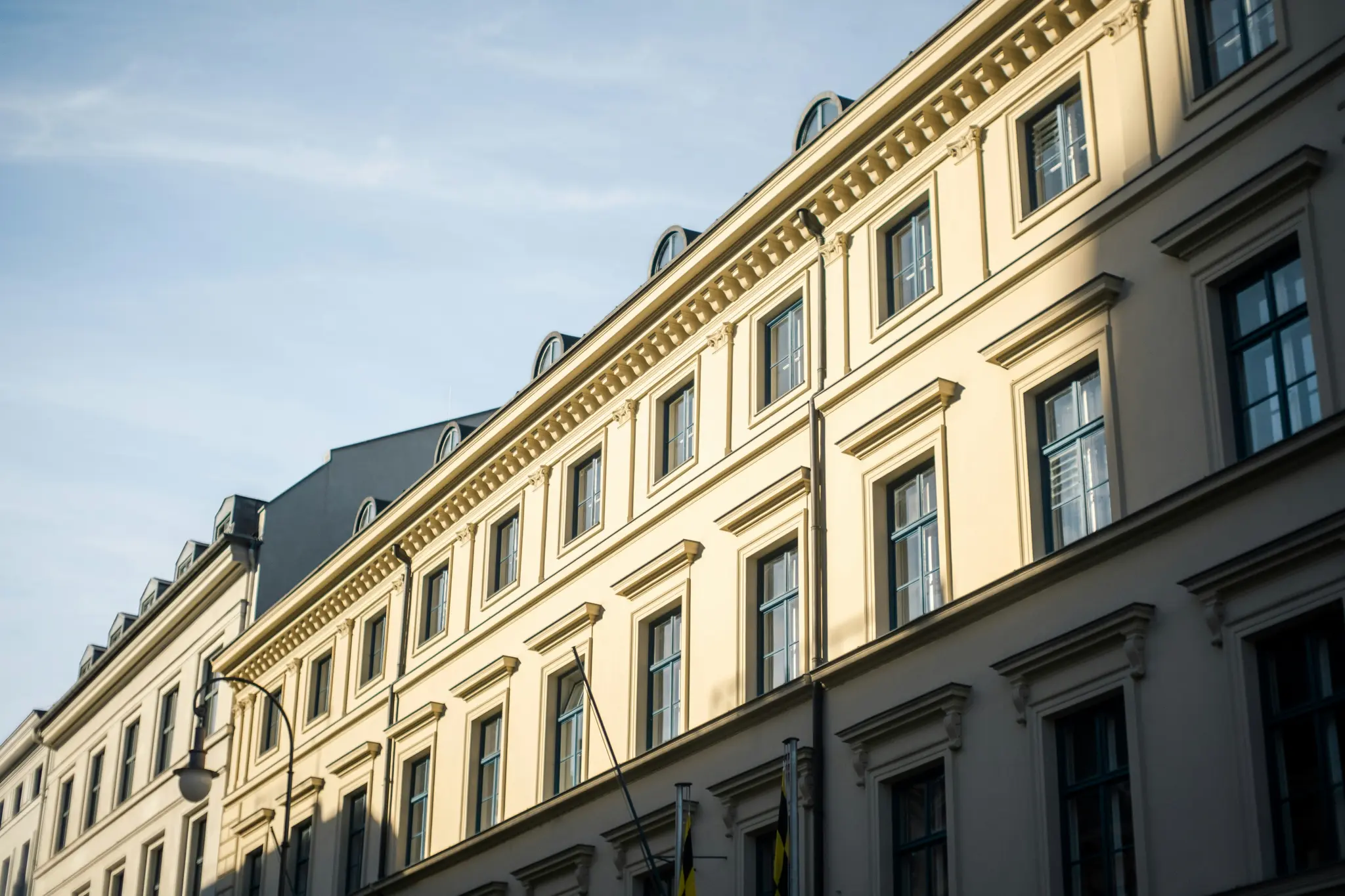 A low-angle view of several cream-colored classical buildings with many windows, under a clear blue sky. Sunlight…