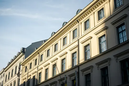 A low-angle view of several cream-colored classical buildings with many windows, under a clear blue sky. Sunlight…