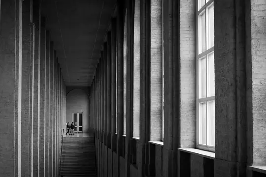 A black and white photo of a long, narrow hallway with numerous tall, rectangular pillars lining both sides. Sunlight…