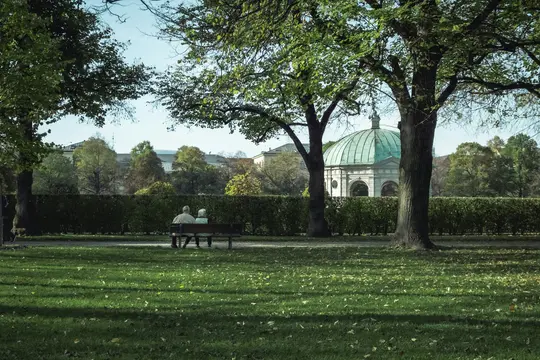 To personer sidder på en parkbænk, ryggen til beskueren, og kigger på en hæk. Bagved er en kuppelbygning og træer synlige.