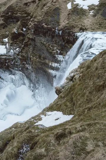 Et kraftfuldt vandfald styrter ned ad en mørk, stenet klippevæg, omgivet af sne og is, ned i et hvidt bassin.