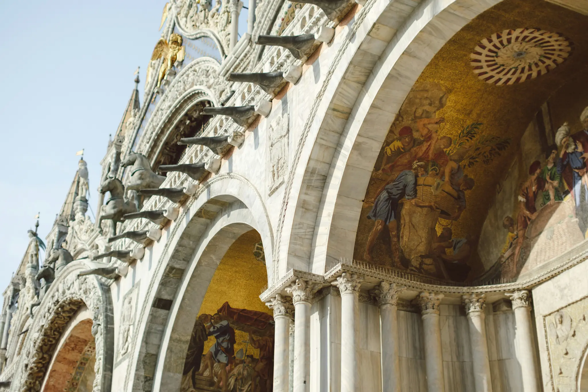 Een opname van onderaf van de sierlijke witmarmeren gevel van de Basiliek van San Marco, met gouden mozaïeken en sculpturen.