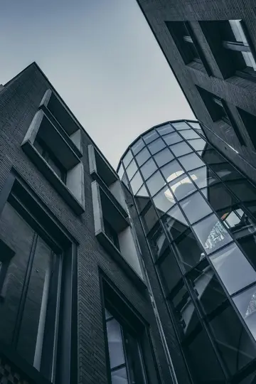 A low-angle view of modern buildings featuring dark brick facades and a central curved glass structure under an overcast sky.