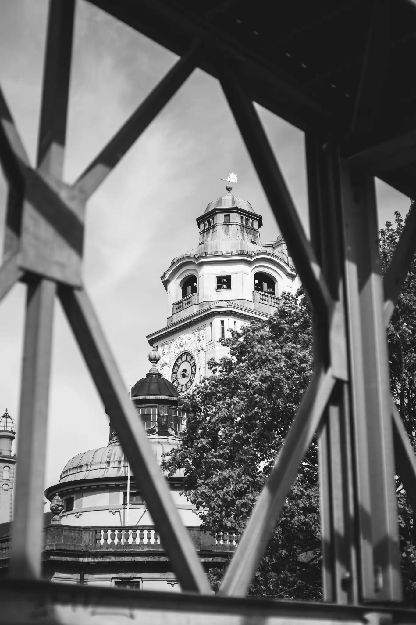 A black and white photo of a grand building with a prominent clock tower, seen through the diagonal bars of a metal…