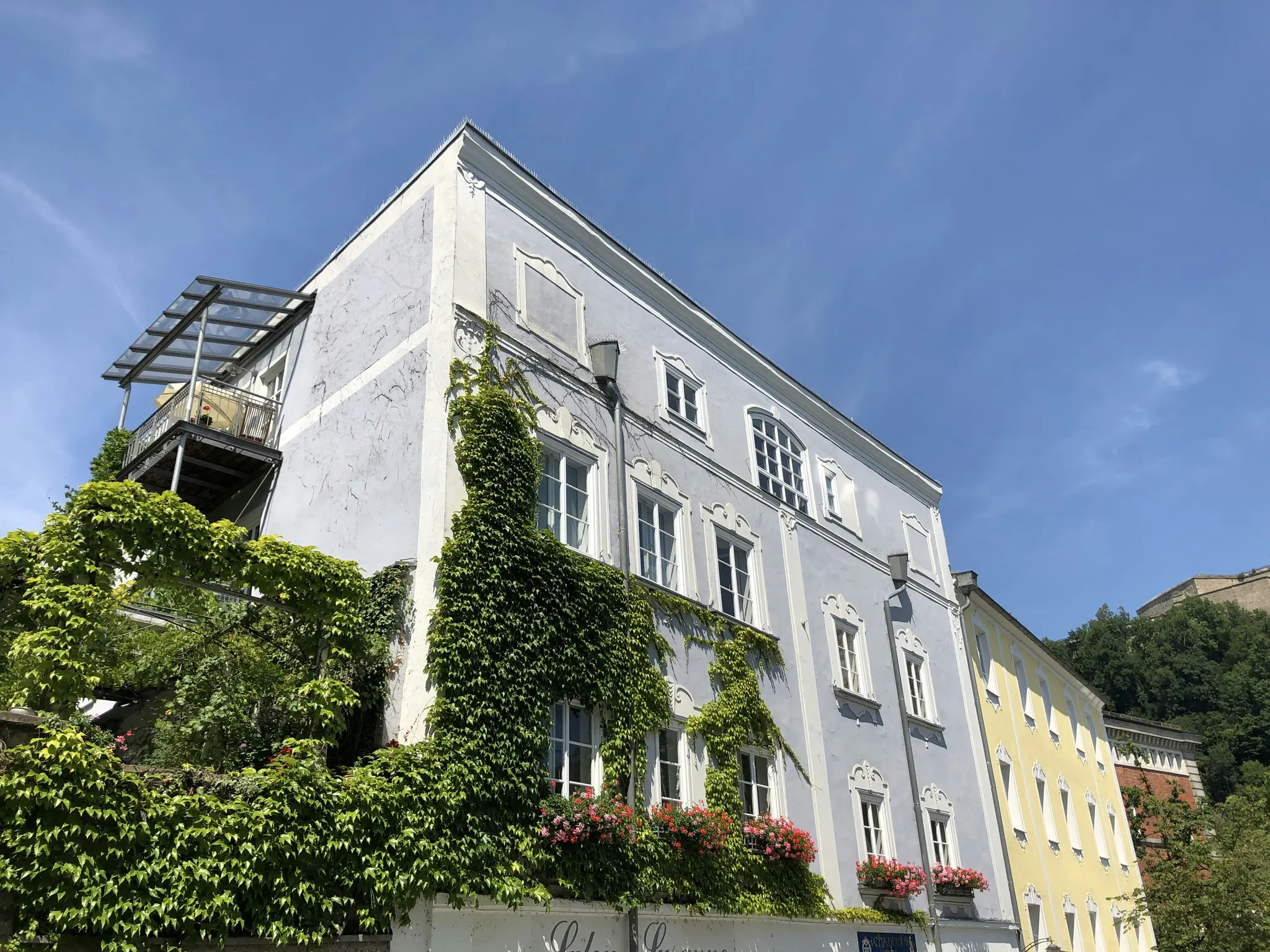 A light blue building with white trim, heavily covered in green ivy and red flowers, stands against a clear blue sky. A…