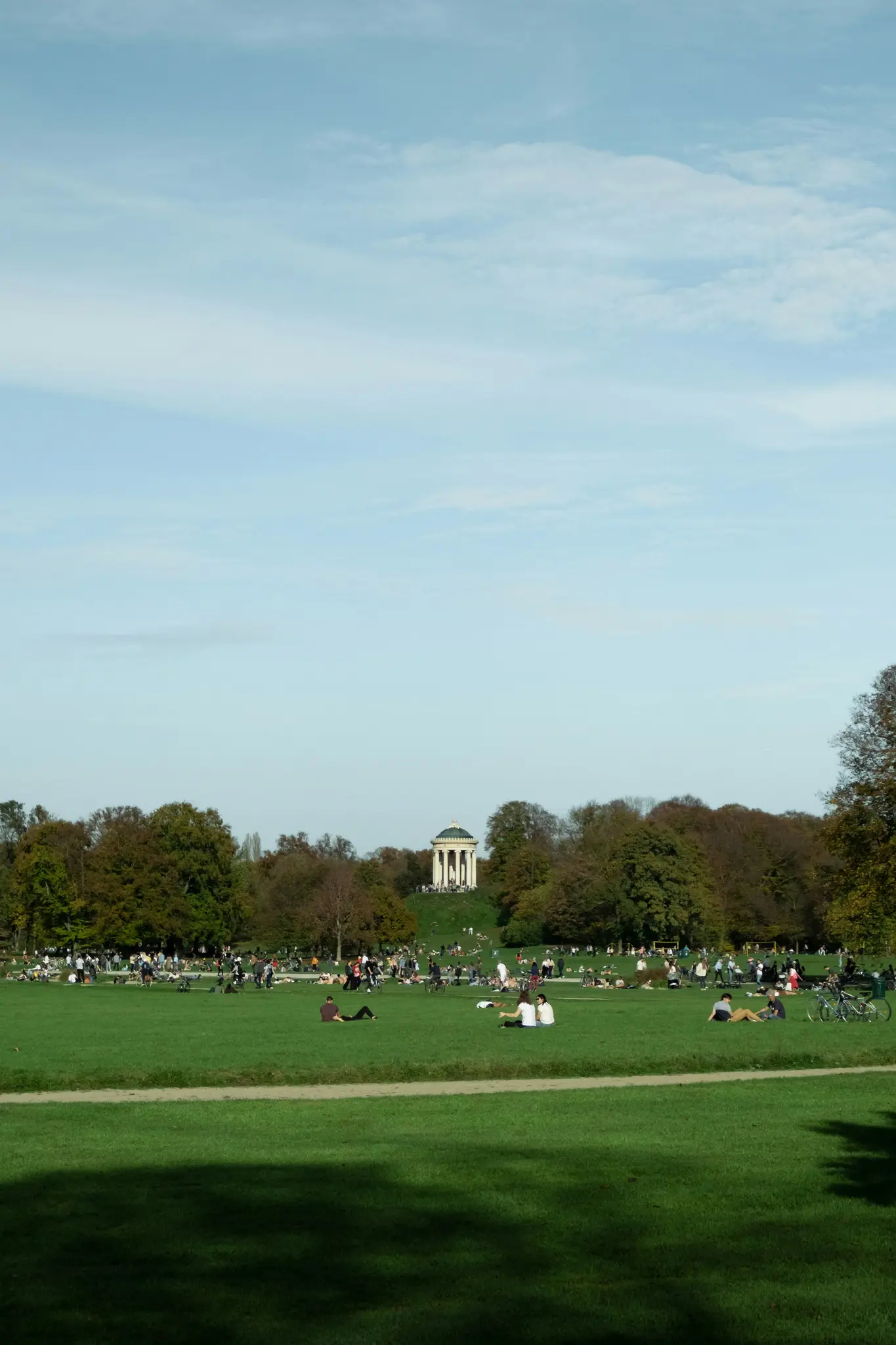 Et bredt landskabsfoto af en park med mange mennesker på grønt græs. Et hvidt klassisk tempel står på en fjern bakke.