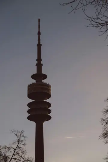 A tall, dark telecommunications tower with multiple observation decks rises against a pale, overcast sky. Bare tree…
