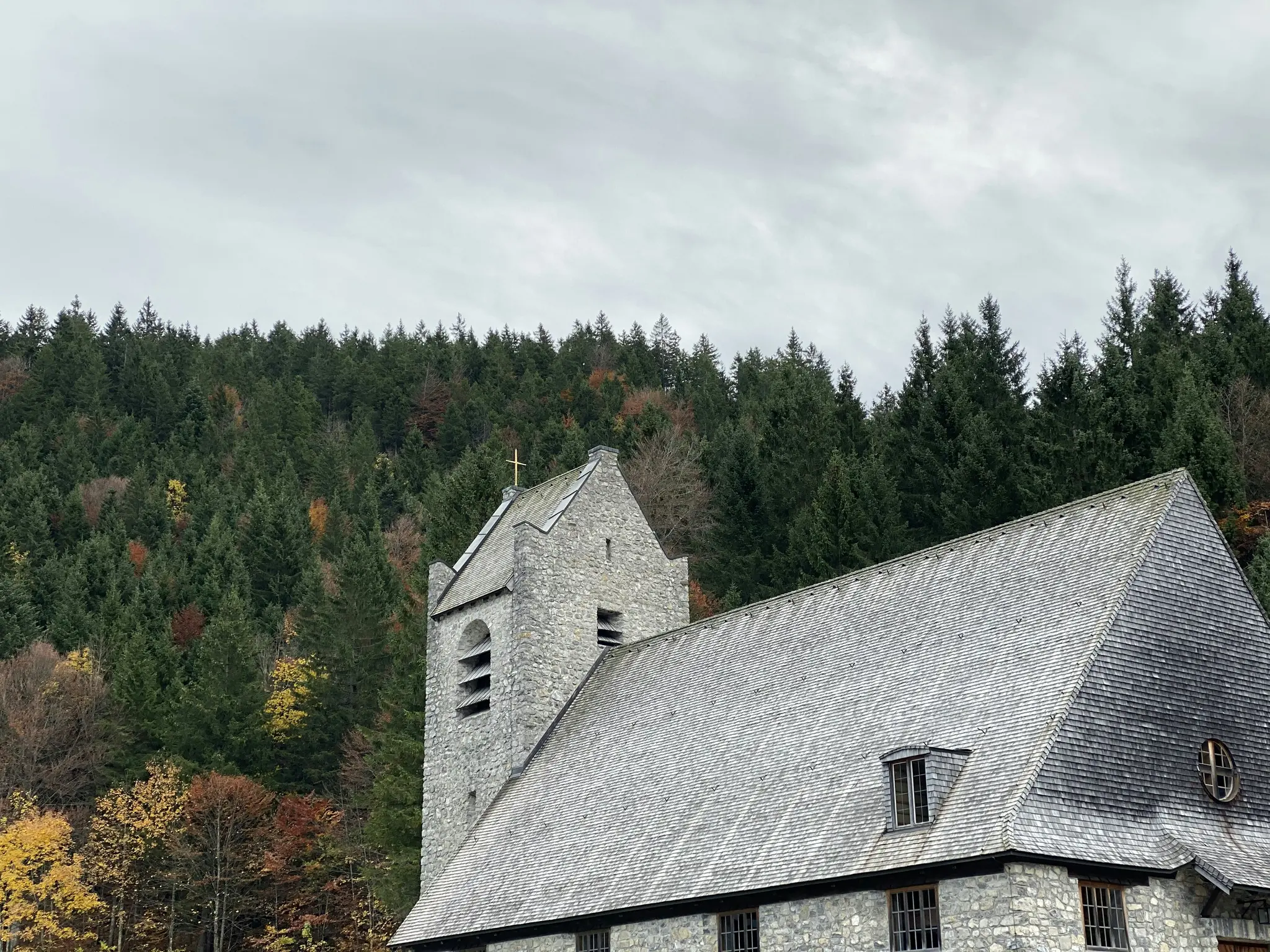 En stenkirke med et gråt skifertag og et klokketårn med et gyldent kors, sat op mod en skov med efterårsfarver.