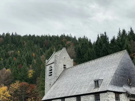 En stenkirke med et gråt skifertag og et klokketårn med et gyldent kors, sat op mod en skov med efterårsfarver.