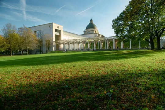 A grand building with a green dome and a modern extension, featuring a long colonnade, overlooks a sunlit grassy park with…