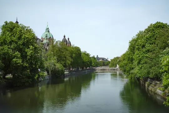 A wide landscape photo of a calm river flanked by dense green trees. Historic buildings with domed roofs are visible.