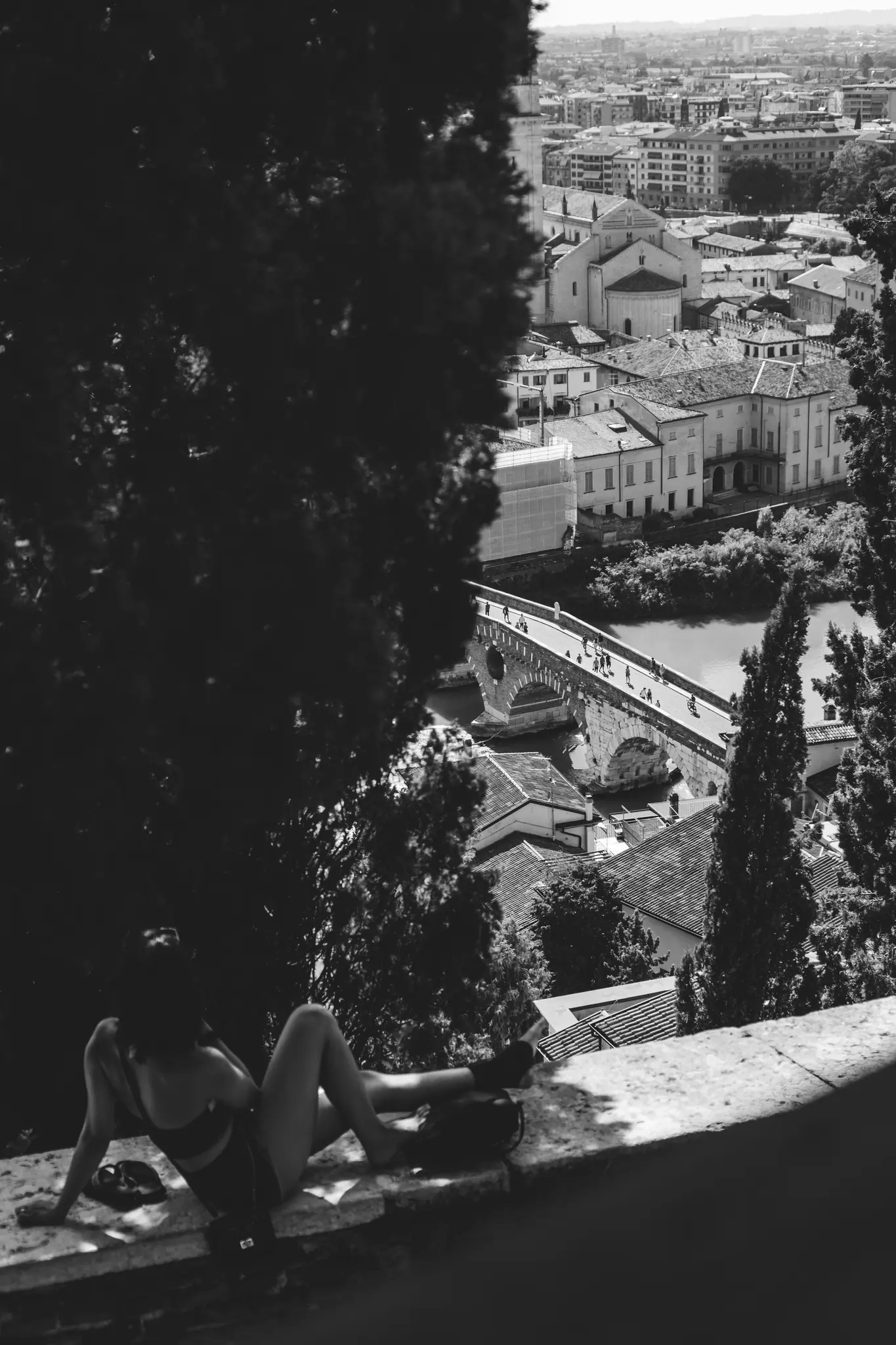 A black and white photo of a person relaxing on a stone wall, looking out over a city with a river and bridge.