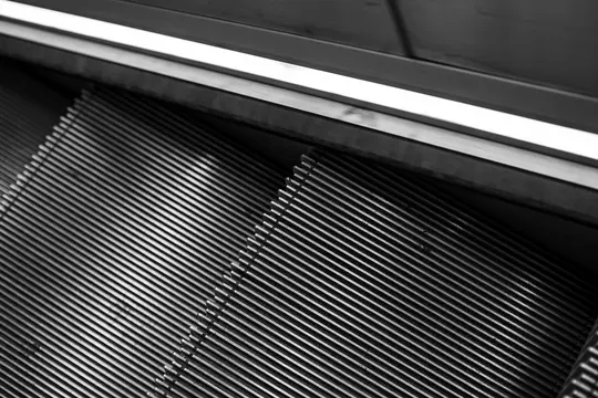 A black and white, high-angle close-up of an escalator, showing the ribbed steps and a smooth handrail.
