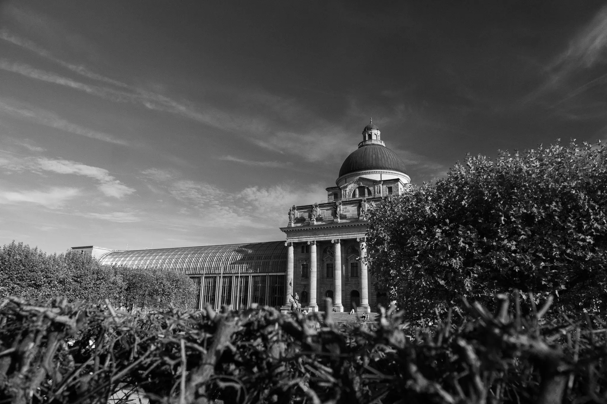 A black and white photo shows a grand domed building with columns and a long glass-roofed extension, seen behind dark…
