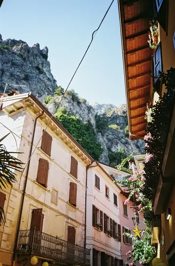 A low-angle view of a narrow street flanked by old buildings with brown shutters, leading to a rocky mountain.