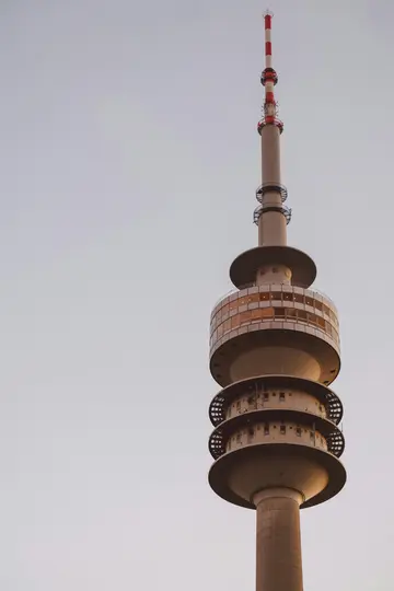 A tall, slender concrete telecommunications tower with a red and white striped antenna at its top, set against a light grey…