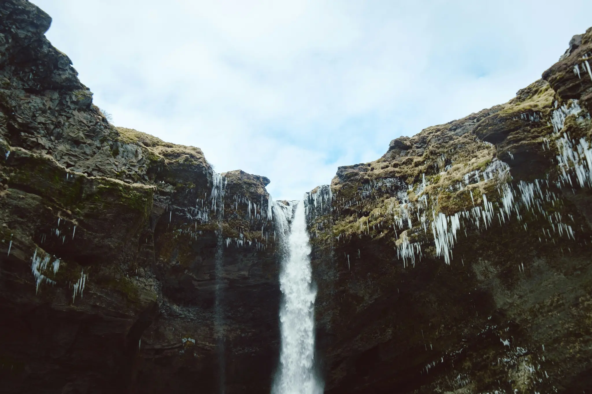 A tall, white waterfall plunges between two dark, rugged cliffs covered in green moss and numerous sharp, white icicles.…
