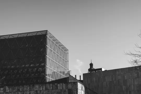 A black and white photo of a modern building with a triangular grid facade next to a stone building with a dome-shaped spire.