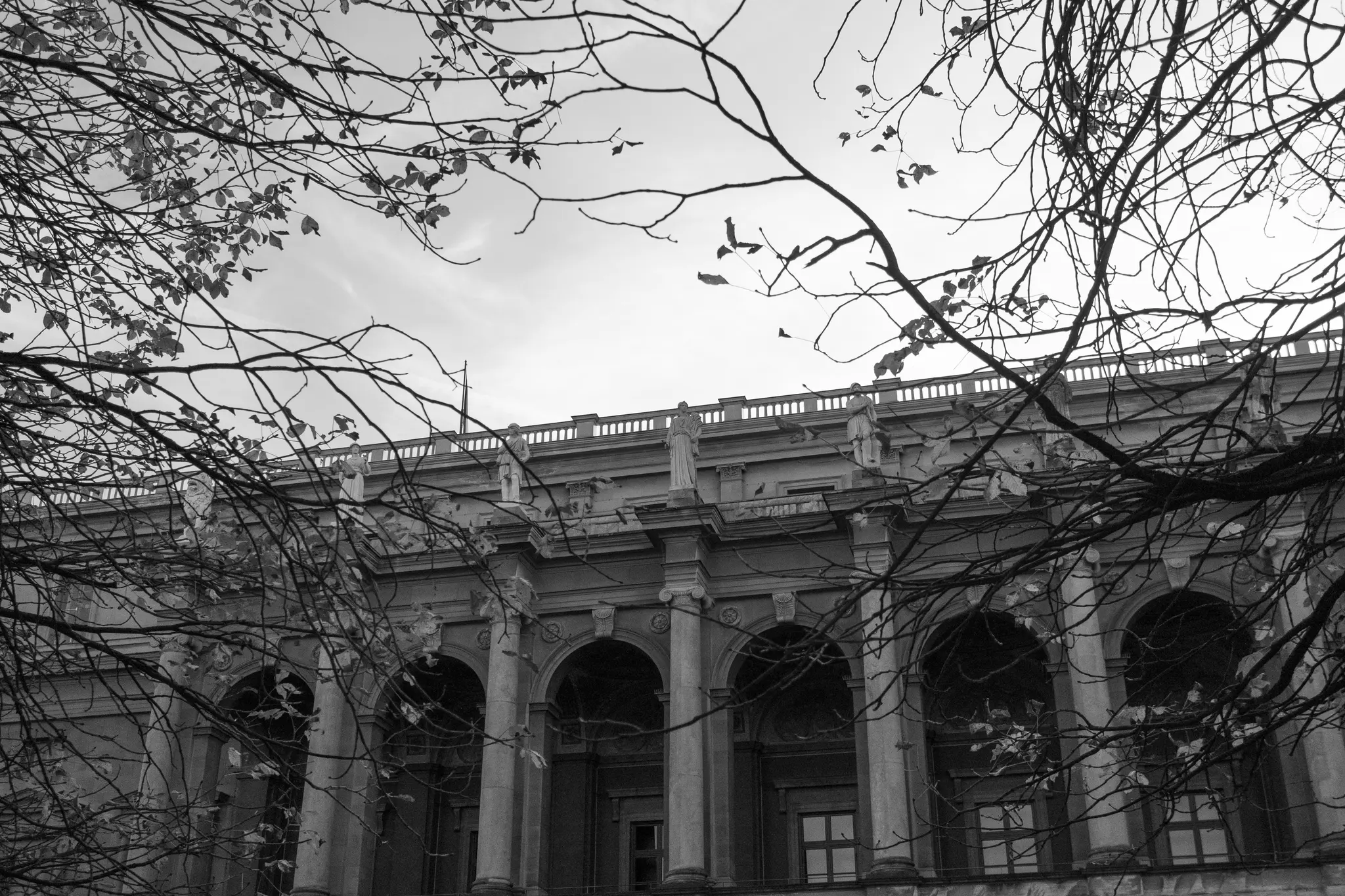 A black and white photo shows a classical building with arched windows and columns, topped with statues and a balustrade.…