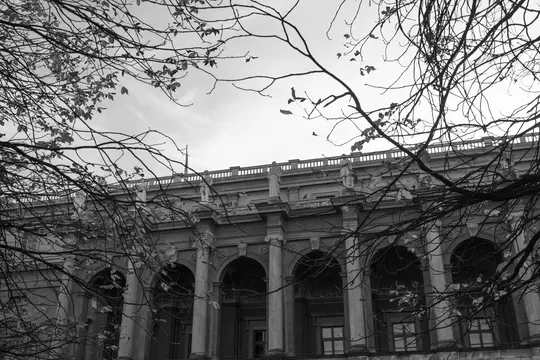 A black and white photo shows a classical building with arched windows and columns, topped with statues and a balustrade.…