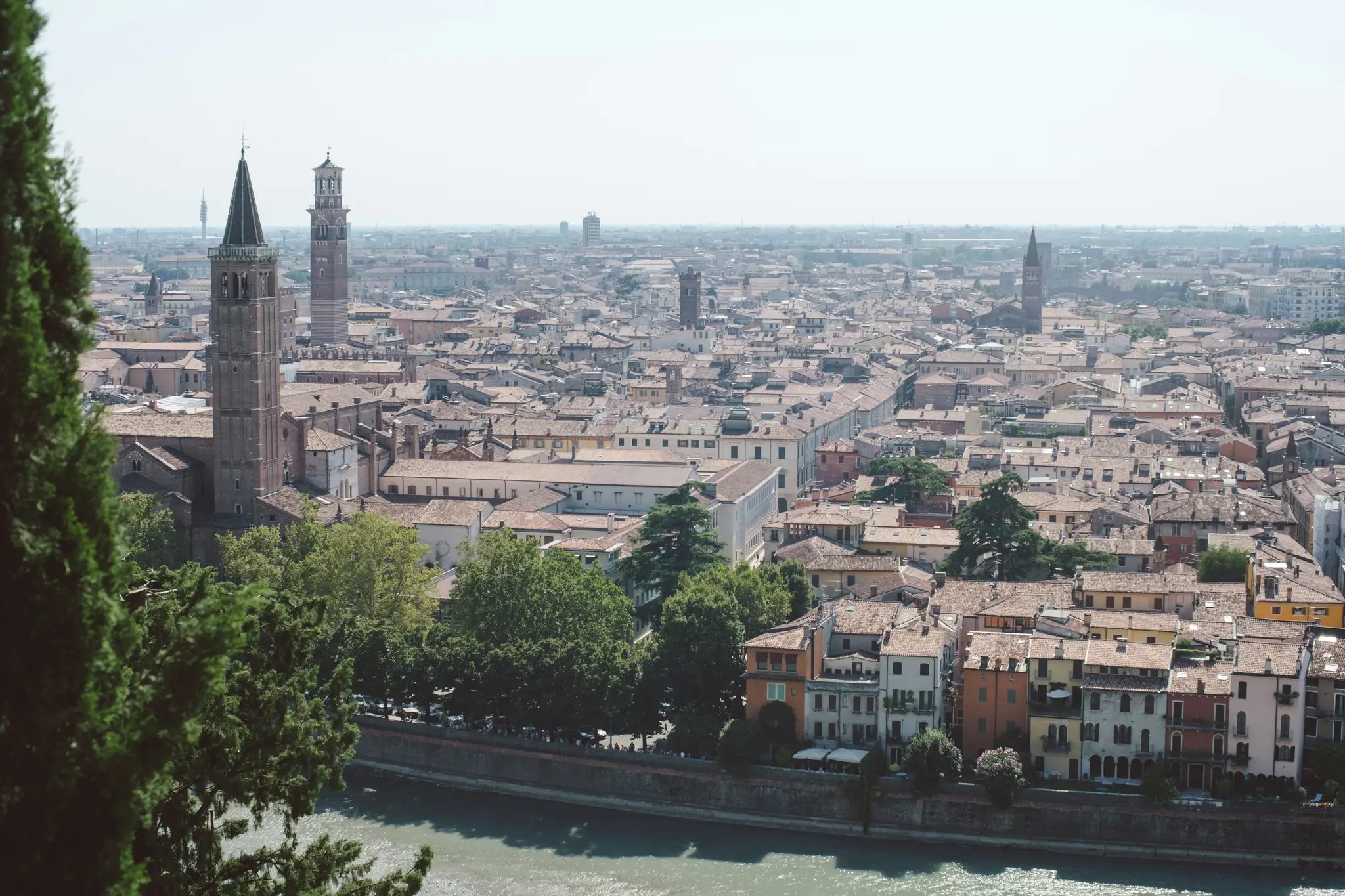 A wide, elevated shot of a city with a river. Terracotta roofs dominate, with two tall bell towers prominent. Green trees…