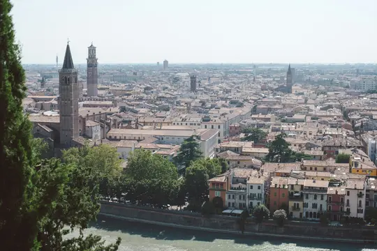 A wide, elevated shot of a city with a river. Terracotta roofs dominate, with two tall bell towers prominent. Green trees…