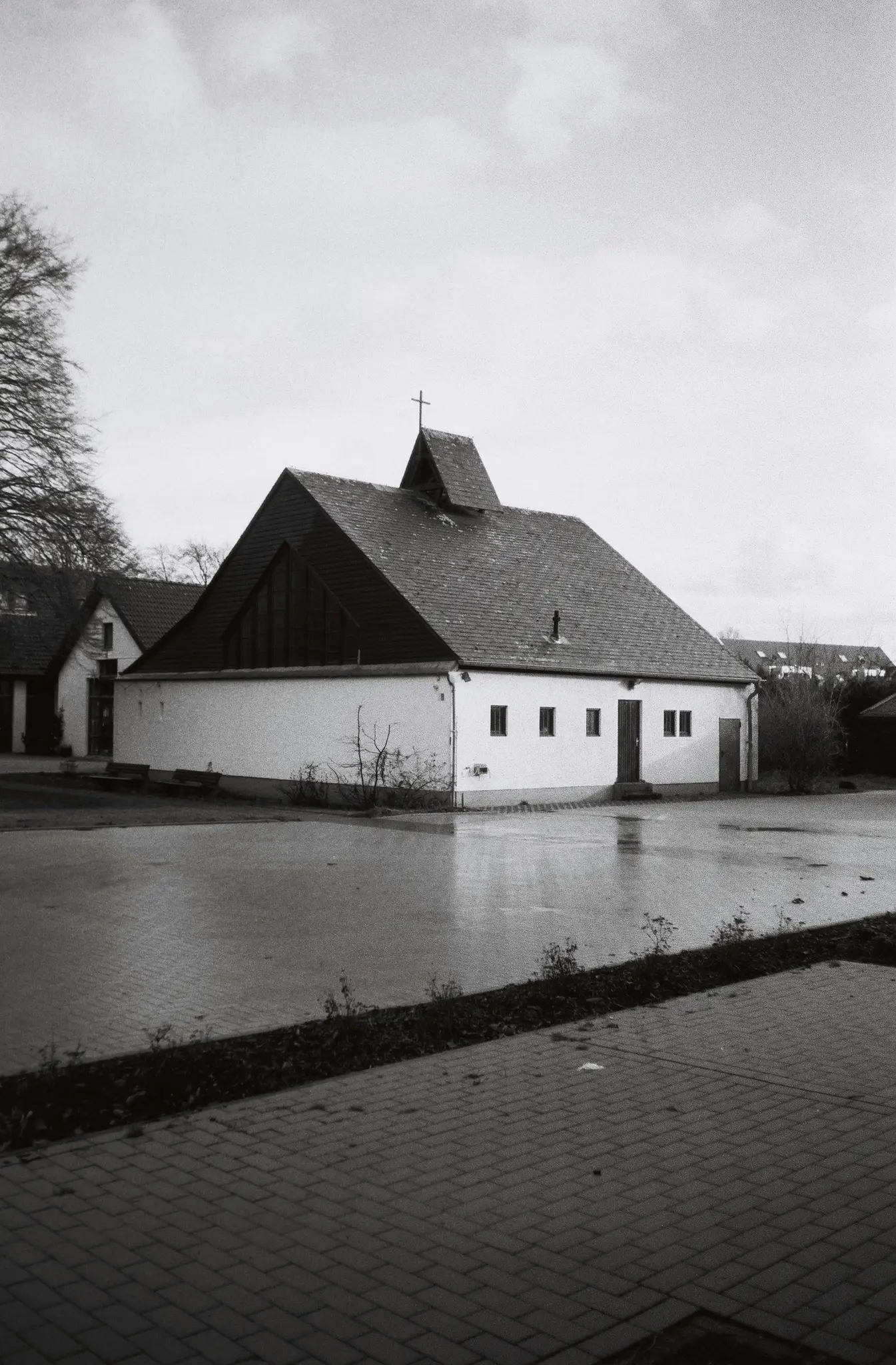 A black and white photo shows a white building with a dark, steeply pitched roof and a small steeple topped with a cross.…