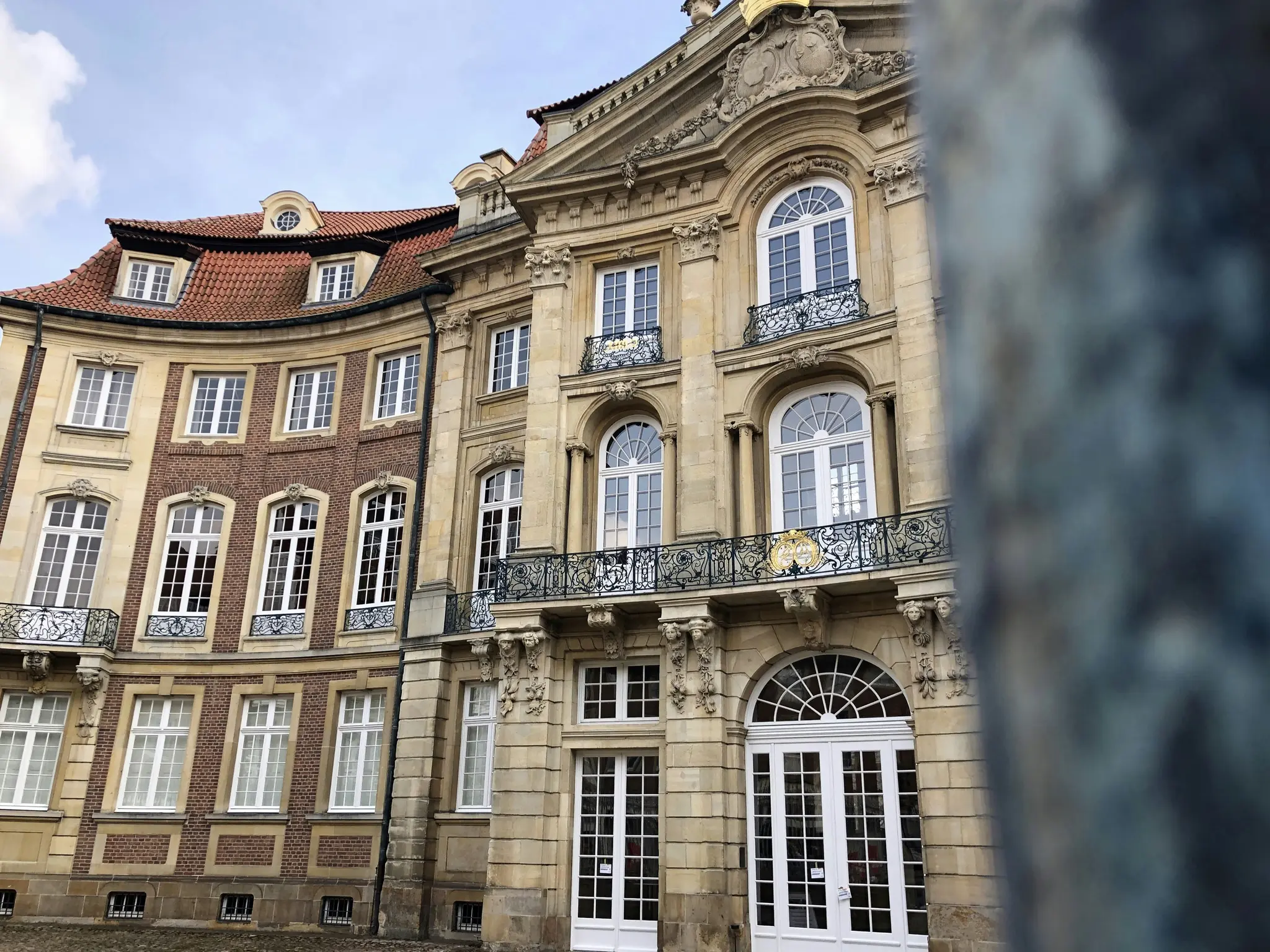 Egy történelmi épület piros cseréptetővel és bézs és tégla homlokzattal, díszes erkélyekkel és íves ablakokkal.