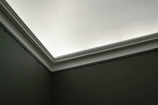 A low-angle shot of a room corner, featuring a dark grey wall and a bright, white illuminated ceiling with molding.