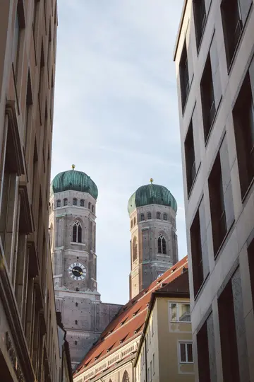To høje, lyse kirketårne med grønne, løgformede kupler mod en bleg blå himmel, indrammet af moderne bygninger.