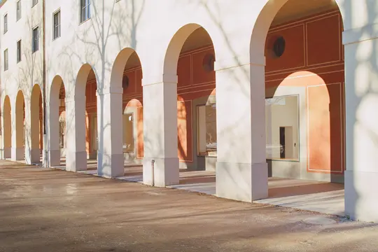 A long perspective view of a cream-colored colonnade with multiple arches and pillars, casting distinct shadows on the…