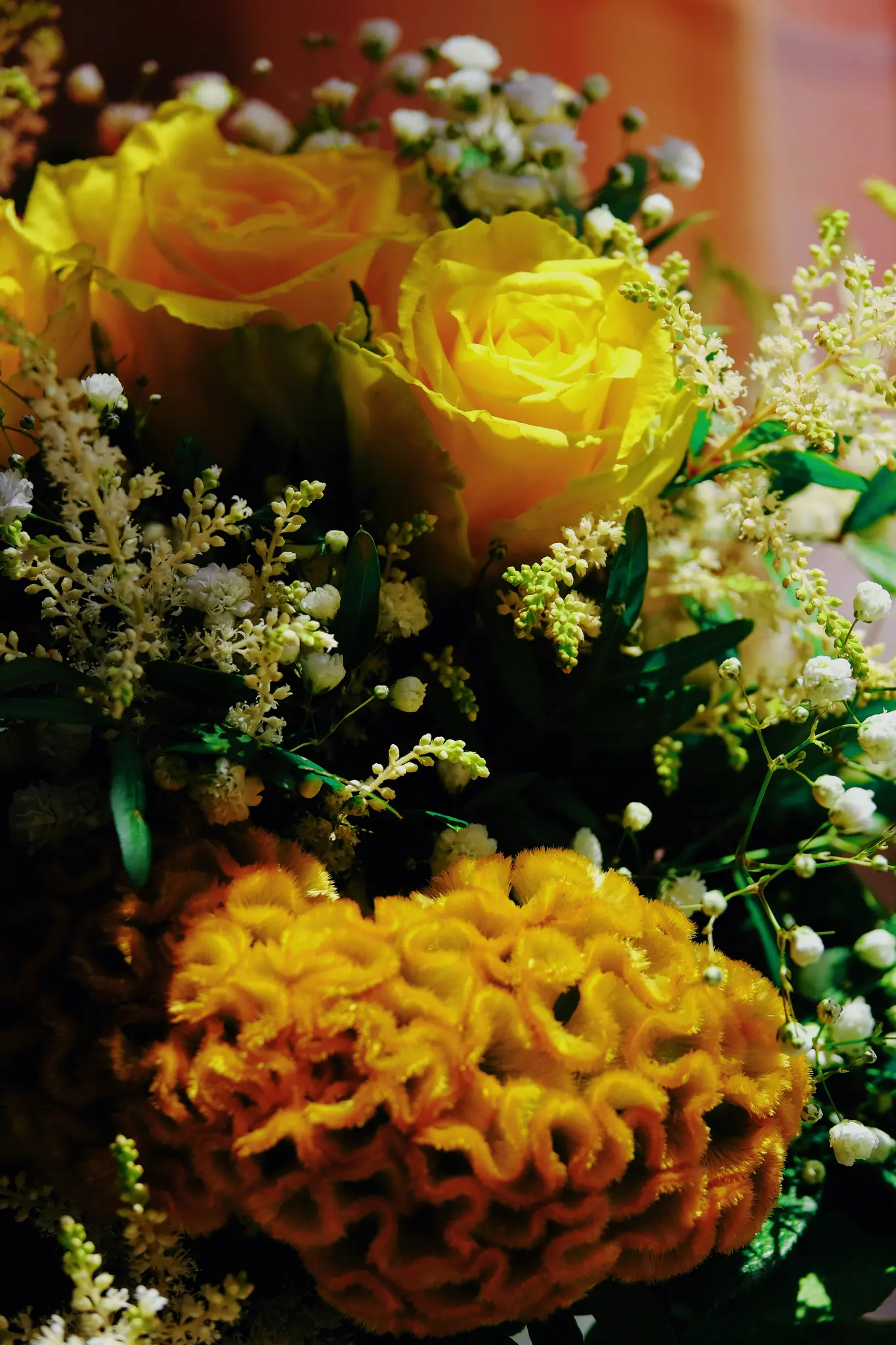 A close-up photo of a bouquet featuring bright yellow roses, white baby's breath, and a large orange cockscomb flower.