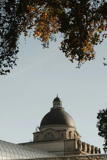 A grand domed building with a smaller cupola on top, framed by dark autumn leaves against a pale blue sky.