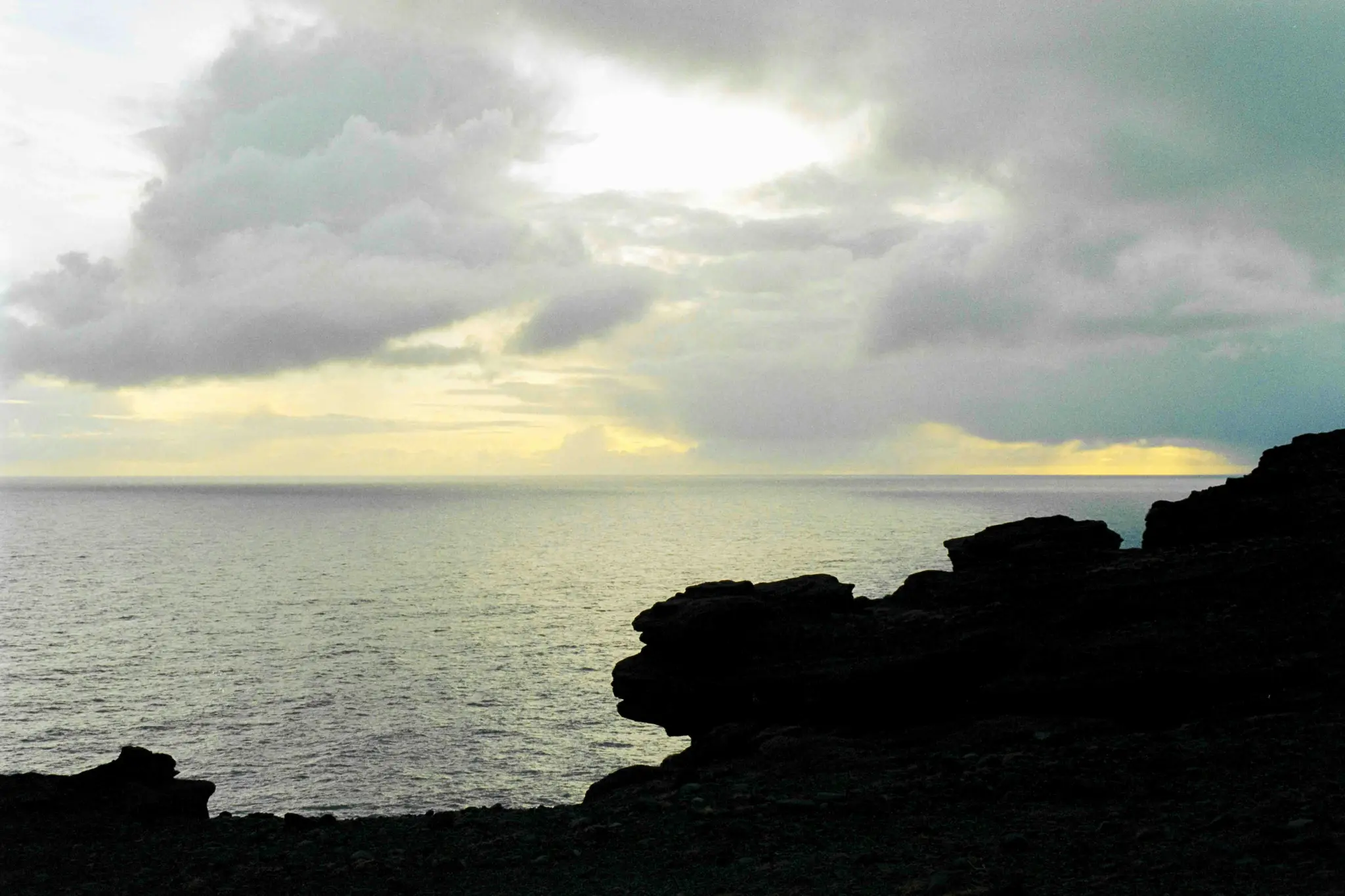 A wide landscape photo of a dark ocean under a cloudy sky, with a bright yellow line on the horizon. Dark cliffs silhouette…