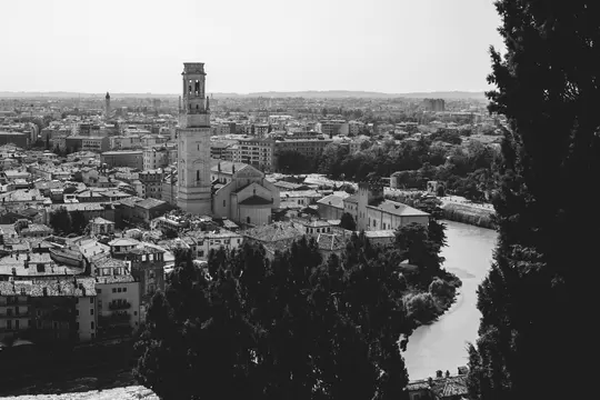 A high-angle black and white photo of a city, featuring a tall bell tower, a river curving through the urban area, and…