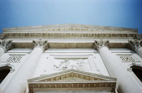 A low-angle shot of a white classical building facade with ornate columns, a pediment, and a carved relief, against a clear…