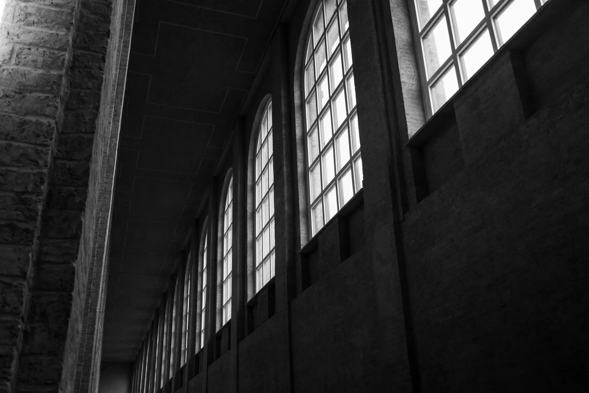 A black and white photo of a long, empty corridor with a row of tall, arched windows on the right and a stone wall on the