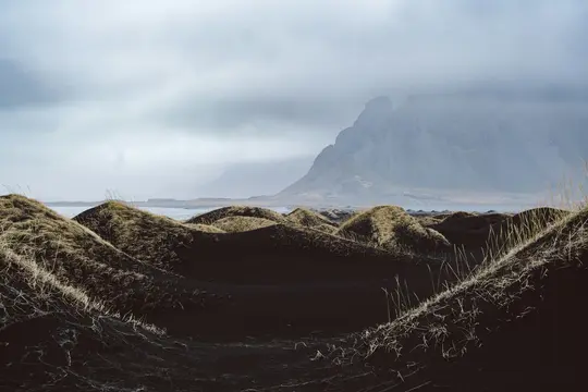 A dramatic landscape features dark sand dunes with patches of dry, light-colored grass in the foreground. A body of water…
