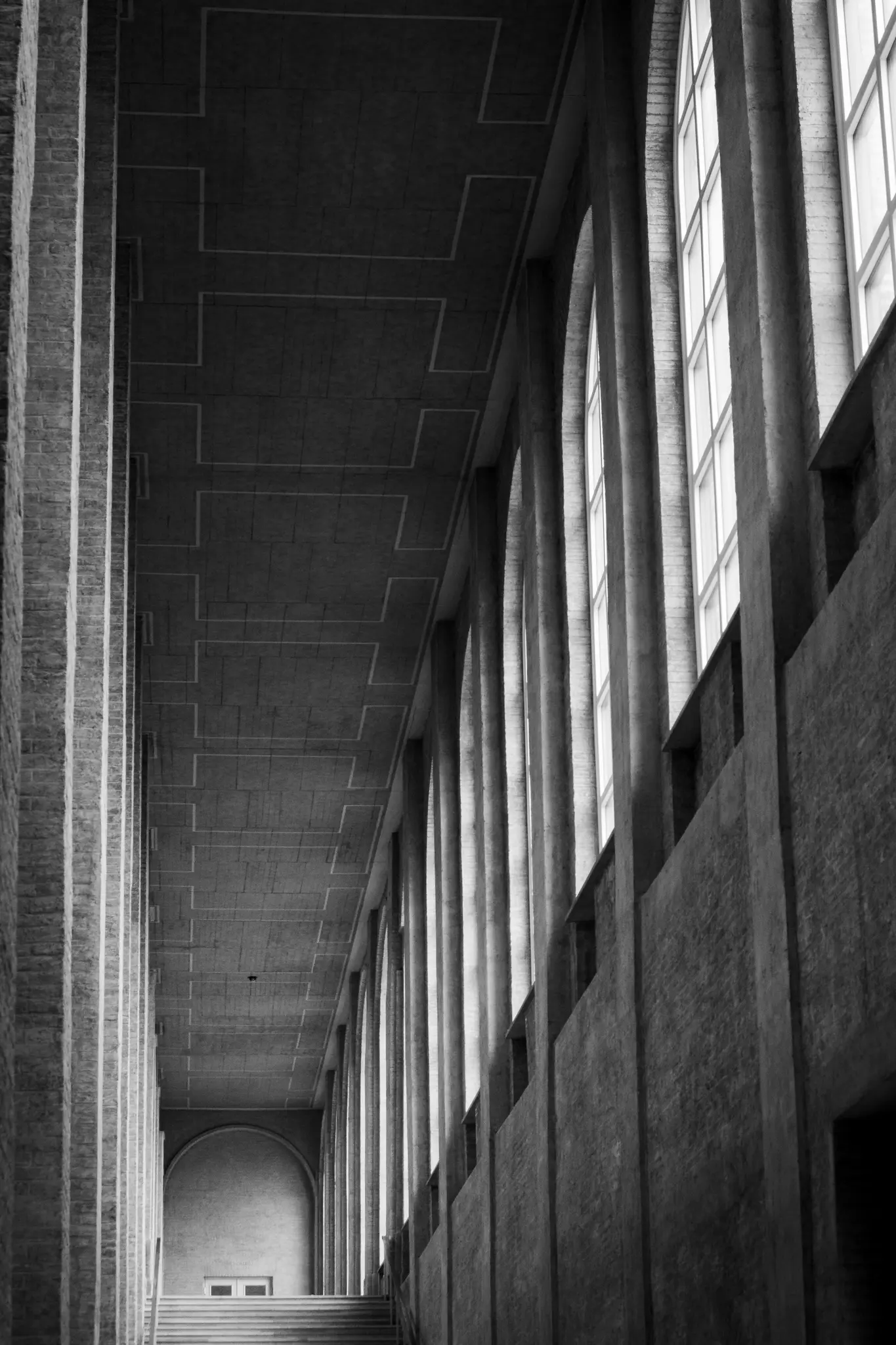 A black and white photo of a long, empty hallway with a high, patterned ceiling and a row of tall arched windows.