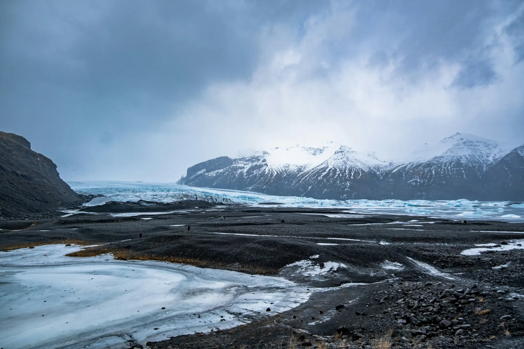 A wide landscape shows a vast glacial plain with dark volcanic rock and patches of ice, leading to a blue glacier and…